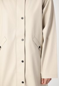 Beige vattentät regnjacka med rak skärning, stora sidofickor, svarta tryckknappar och synliga sömdetaljer. Mjuk textur.