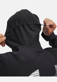 Schwarze Jacke mit Kapuze, ausgestattet mit einem Zugmechanismus. Strukturierter Stoff mit reflektierenden Akzenten auf dem Rücken, die das Markenlogo hervorheben.