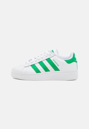 Baskets Adidas Superstar blanches avec des rayures vertes, languette et logo au talon, dotées de lacets à l'avant et d'une coque en caoutchouc à l'avant.