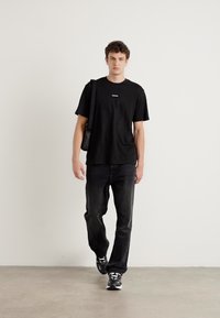 Black cotton t-shirt with small logo, paired with black jeans. Model carries a black bag and wears black athletic shoes. Minimalist design.
