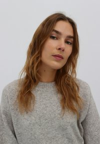 Hellgrauer, gestrickter Pullover mit einer strukturierten Oberfläche und einem runden Ausschnitt. Das Model hat welliges, schulterlanges Haar und einen neutralen Hintergrund.