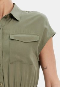 Abito camicia corto a maniche corte di colore verde oliva con colletto a punta e due tasche a flap sul petto, realizzato in tessuto liscio.