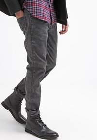 Šedé džínové slim fit kalhoty s jemnou texturou, doplněné černými koženými kotníkovými botami na tkaničky. Oblečení zahrnuje červenou a modrou károvanou košili.