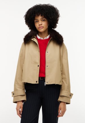Ung kvinde iført en beige croppet jakke med pelskrave, rød sweater, hvid skjorte og sorte bukser med nålestriber, stående mod hvid baggrund.