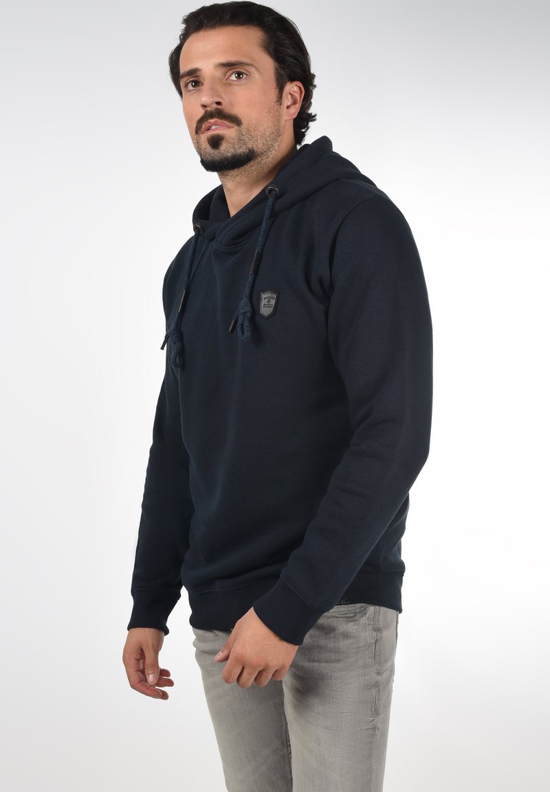Marineblauwe hoodie met koordjes, ribgebreide manchet en een klein logo-patch op de borst. Het materiaal lijkt zacht en gestructureerd.