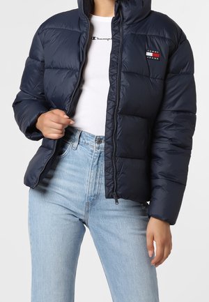 Person iført en åbnet navy dunjakke over en hvid Champion t-shirt og lyseblå højtaljede jeans mod en ensfarvet baggrund.