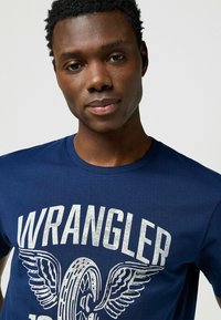 T-shirt in cotone blu navy con una grande stampa bianca "WRANGLER" invecchiata, completa di dettagli grafici di ali e pneumatici sul davanti.
