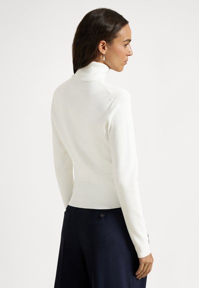 Žena v boku otočená, oblečená v bielom roláku a tmavomodrých nohaviciach na bielom pozadí.