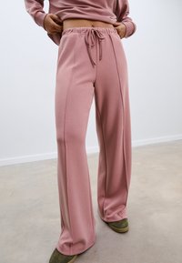 Ružičaste široke hlače od mekanog materijala s remenom za podešavanje u struku, sa ravnim šavovima i opuštenim krojem. Uparene s zelenim tenisicama.