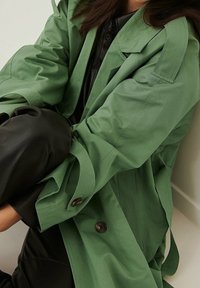 Kobieta w zielonym trenczu i czarnych spodniach, siedząca z ramionami objętymi wokół kolan przy jasnej ścianie.