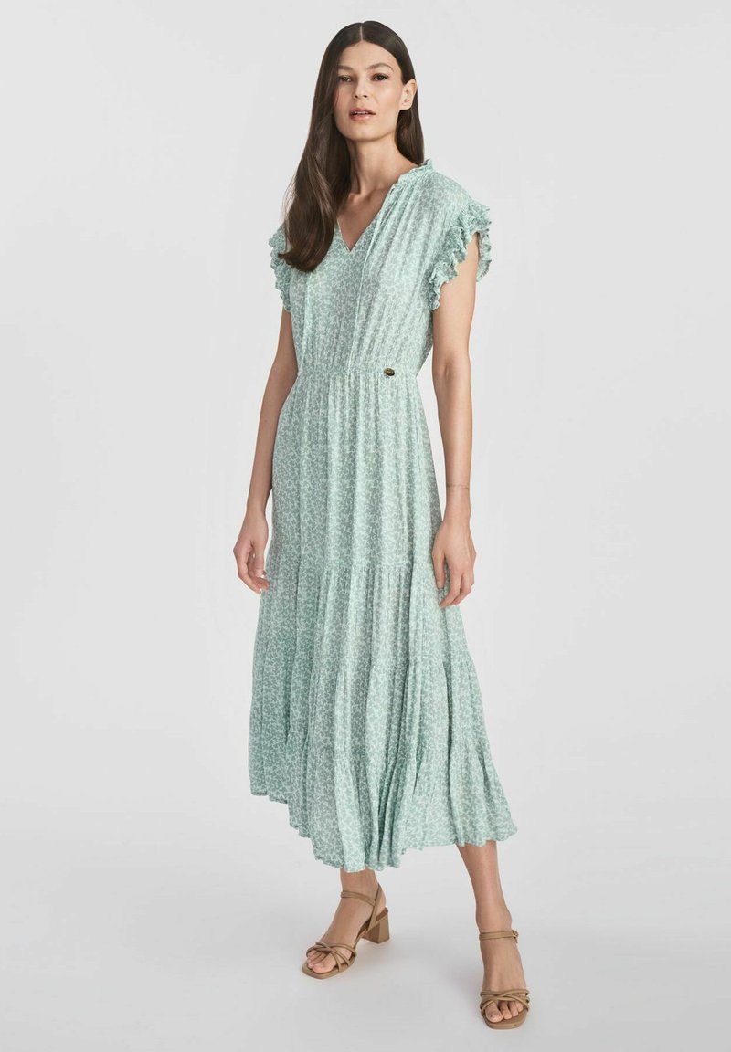 Jasnozielona sukienka maxi w kwiaty z falbankowymi rękawami, gumowym pasem, warstwową spódnicą i małym logo. W połączeniu z beżowymi sandałami na obcasie.