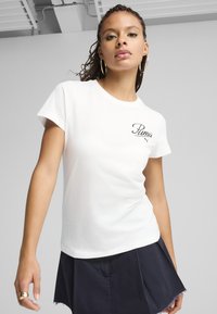 Witte katoenen t-shirt met korte mouwen, voorzien van geborduurd "Puma" logo in het zwart. Model in een zwarte rok staat tegen een lichte achtergrond.