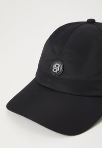 Svart caps laget av glatt stoff med en avrundet krone og en buet skygge. Har et sirkulært logo på fronten.