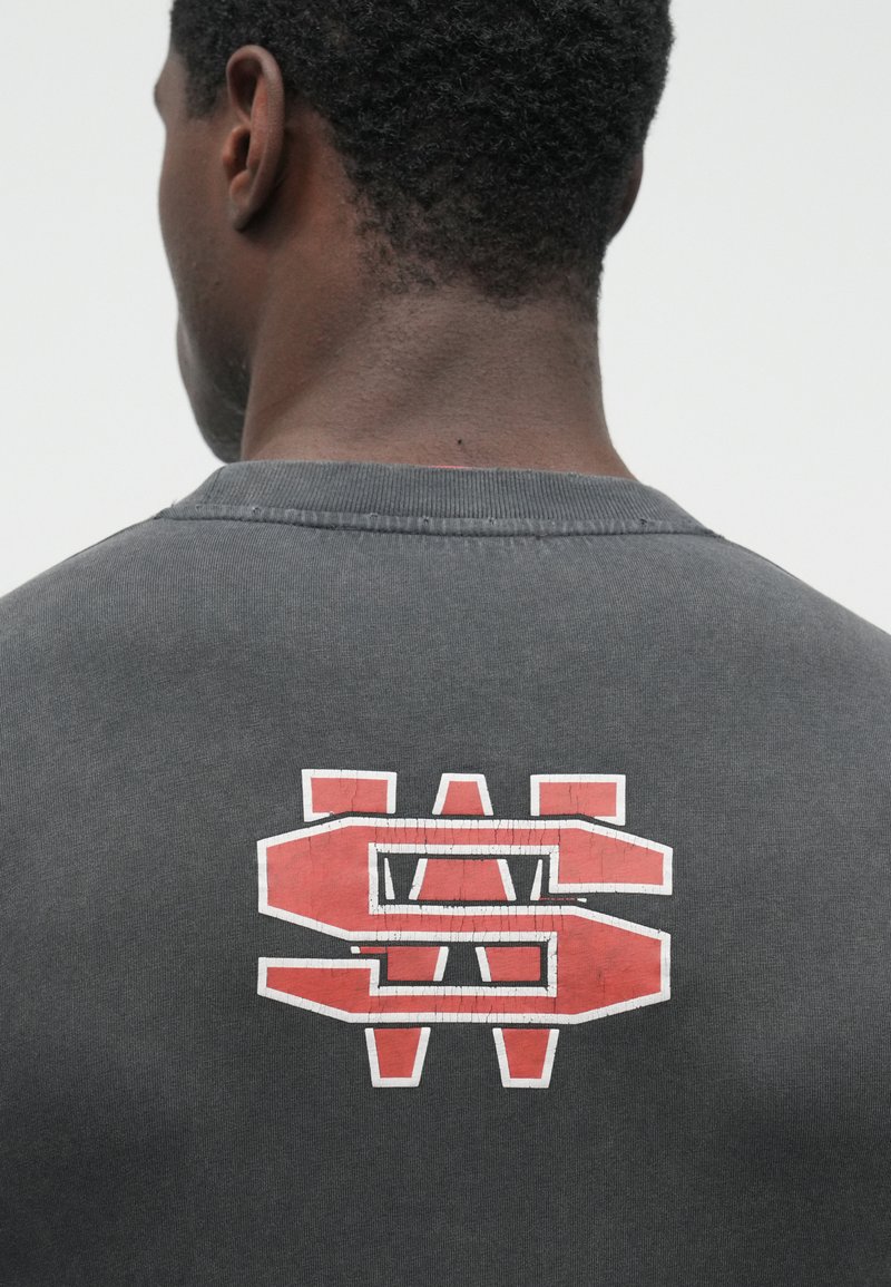 Bagfra view af en person iført en mørkegrå skjorte med store revnede røde og hvide stiliserede bogstaver "WS" på den øverste del af ryggen.