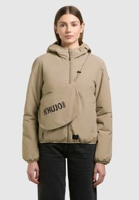 Jeune femme portant une doudoune beige à capuche avec une pochette bandoulière étiquetée "KHUJO 2001" et un pantalon noir, debout devant un fond uni.
