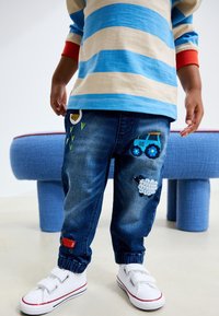 Denimbukser med blå traktor- og fårebroderi, elastiske manchetter og en glat tekstur. Kombineret med hvide sneakers med røde accenter.