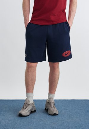 Hombre con pantalones cortos deportivos azul marino con logo rojo de Nike, camiseta roja, calcetas grises y zapatillas Nike grises, de pie sobre una alfombra azul.