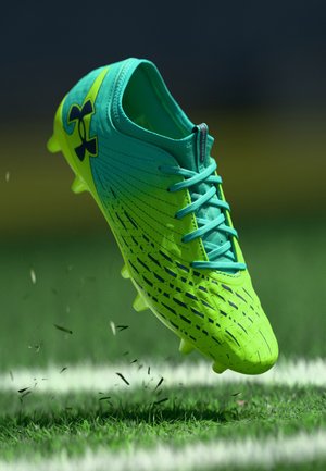 Taco de fútbol en verde y aqua con superficie texturizada, diseño con cordones y tacos debajo, con un logotipo negro y un acabado degradado.