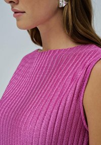 Primer plano de una persona que lleva una camiseta sin mangas de punto en color rosa y un gran arete plateado, con el cabello largo y castaño visible.