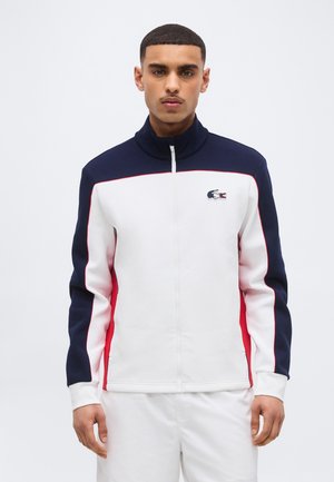 Mees seisab näoga ettepoole, kandes valget jope, millel on mereväe- ja punased värvilahendused ning väikese krokodillilogoga rinnal.