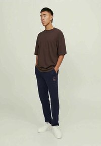 Young man stands with hands in navy sweatpants pockets, wearing a loose brown T-shirt and white sneakers against plain background.