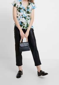 Blusa floreale a maniche corte in bianco, verde e blu; pantaloni in finta pelle nera; piccola borsa nera con manico circolare in legno; scarpe nere.