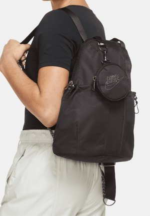 Person iført sort t-shirt og hvide bukser, der bærer en sort Nike rygsæk med en rund Nike pung fastgjort på bagsiden.