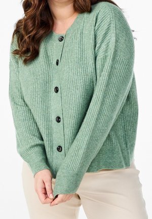 Woman wearing a ribbed, mint green buttoned cardigan with black buttons and beige pants, hands clasped in front.