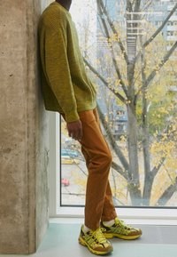 Pull tricot vert olive, pantalon orange brûlé et chaussures de course multicolores. Debout contre un mur en béton avec une fenêtre en arrière-plan.