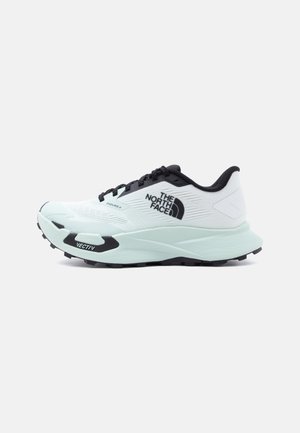Zapatilla de trail running en blanco y negro con el logo de "The North Face", cordones negros y suela gruesa y estriada etiquetada como "VECTIV".