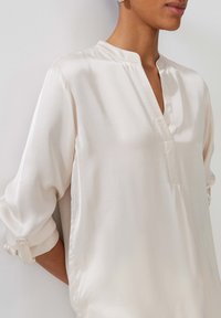 Blusa di seta color crema con colletto alla cinese, maniche lunghe arrotolate, con patta frontale e un leggero brillante.