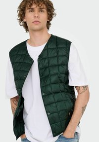 Gilet vert rembourré avec un coussinage en carreaux, boutons-pression et une texture légère, porté sur un t-shirt blanc, présentant des tatouages visibles sur les bras.