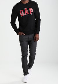 Sudadera negra con el logo rojo de "GAP", de mangas largas, combinada con pantalones gris oscuro y zapatillas blancas. Fondo liso, enfocado en los detalles del conjunto.