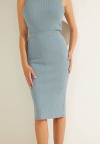 Žena v světle modrém úpletu bez rukávů a odpovídající přiléhavé midi sukni na jednobarevném pozadí.