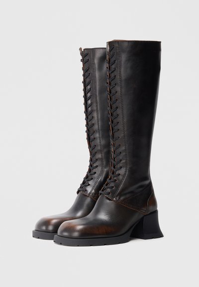 Fekete bőrből készült térdig érő csizma, az elején fűzővel díszítve, kerek orral és vastag sarokkal. Sima felületű és strapabíró talppal rendelkezik.