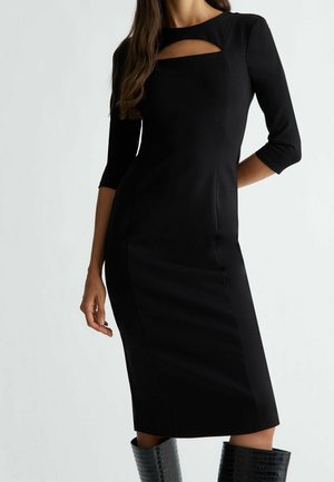 Kobieta ubrana w dopasowaną czarną sukienkę z rękawami do łokcia i prostokątnym wycięciem przy dekolcie, w połączeniu z czarnymi, teksturowanymi kozakami do kolan.