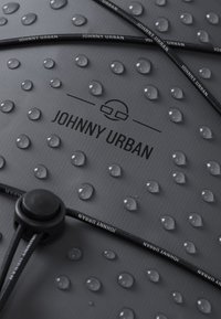 Material grigio con superficie testurizzata caratterizzata da punti rialzati. Gocce d'acqua sulla superficie. Corda nera con testo stampato: "JOHNNY URBAN."