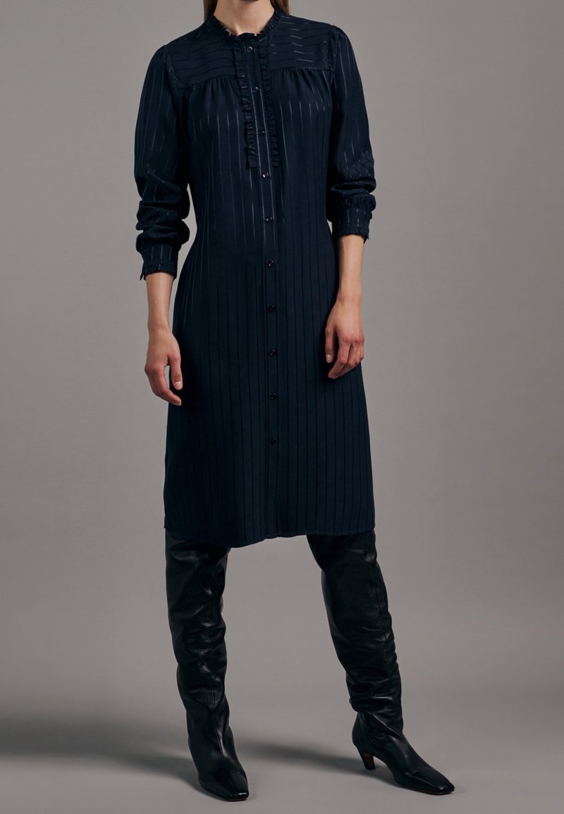 Lång marinblå klänning med vertikala ränder, knapplist framtill och volangärmar. Matchas med åtsittande svarta knähöga stövlar.