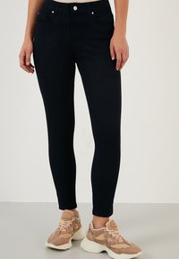 Jeans skinny azul marino de mezclilla, con bolsillos frontales, pasadores para cinturón y una textura sutil, combinados con zapatillas de color claro.