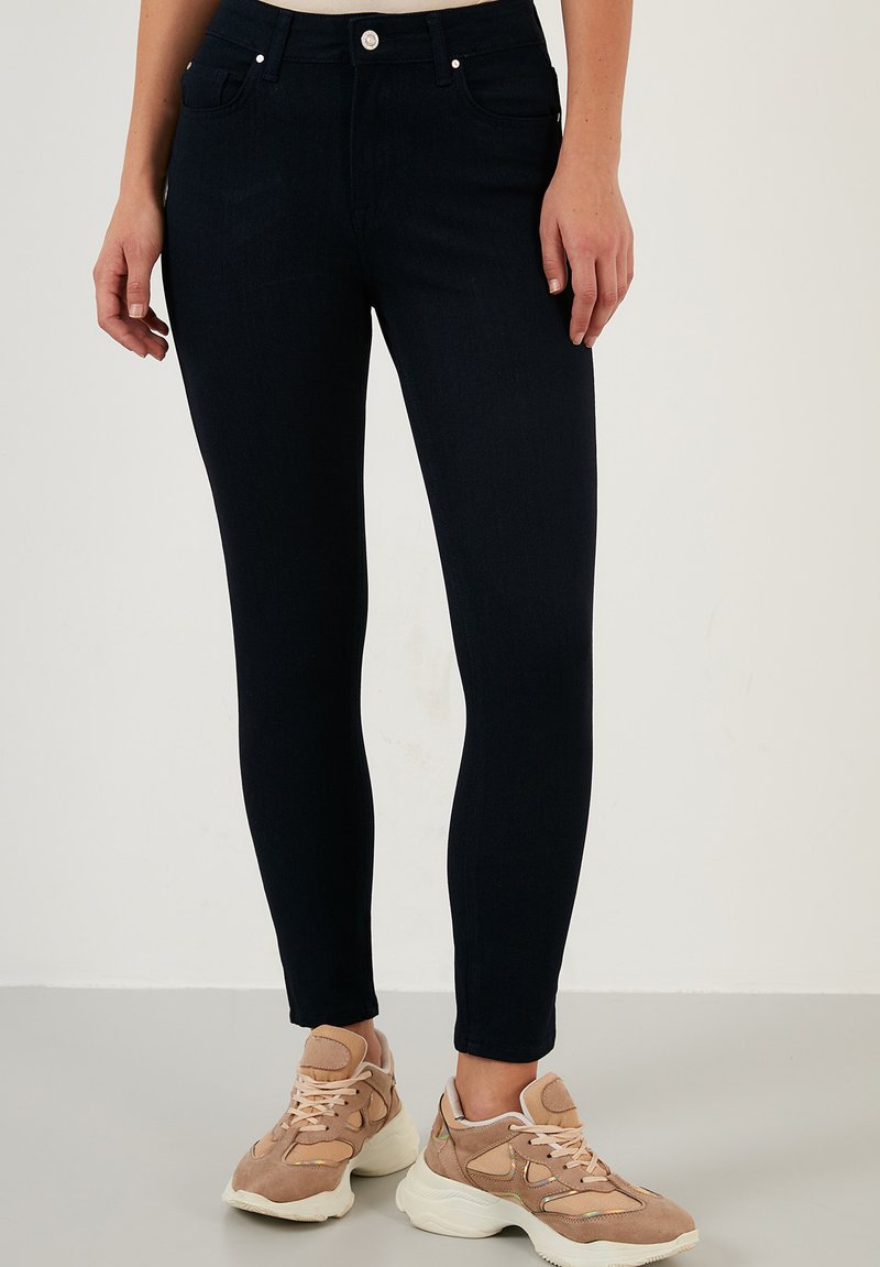 Jeans skinny azul marino de mezclilla, con bolsillos frontales, pasadores para cinturón y una textura sutil, combinados con zapatillas de color claro.