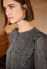 Jeune femme aux cheveux courts bruns, portant un chemisier noir et gris à imprimé léopard avec des détails volantés sur les épaules.