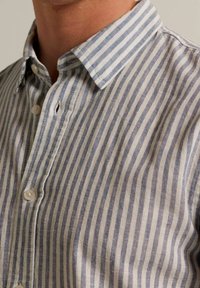 Primer plano de una persona que lleva una camisa de botón a rayas verticales en azul y blanco con cuello puntiagudo.