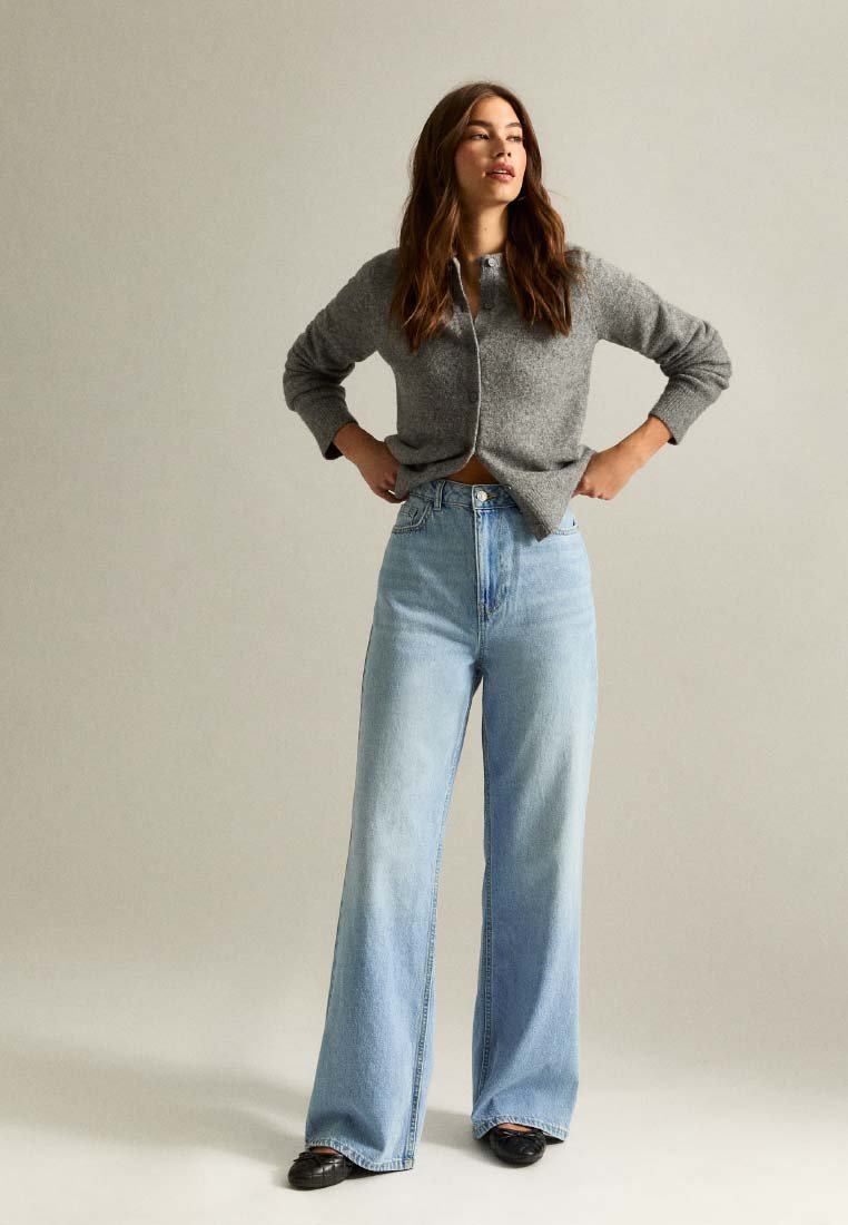 Szary kardigan zapinany na guziki o prążkowanej fakturze, w połączeniu z jasnoniebieskimi dżinsami o wysokim stanie i szerokich nogawkach. Obuwie to czarne, płaskie, błyszczące buty.