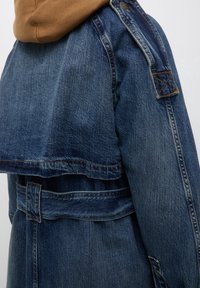 Veste en denim à coupe courte, de couleur bleue, avec une taille ceinturée. Elle présente un dos superposé, des coutures apparentes et des épaulettes.