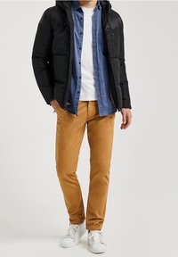 Black puffer jacket, blue button-up shirt, white T-shirt, tan slim-fit pants, white sneakers. Smooth fabric, modern cut, casual style.