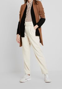 Brown long cardigan with black knit cuffs over a striped shirt, white high-waisted trousers, and white sneakers. Smooth fabrics, tailored fit.