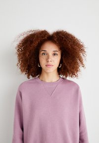 Lyse lilla sweatshirt med rund hals og ribbede detaljer. Modellen har krøllete rødt hår og bruker hoop-øredobber. Enkelt bakgrunn.