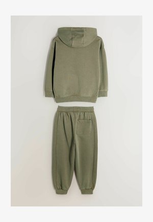 Zaļš kokvilnas kapuce ar aukliņu un ribotām aprocēm, apvienots ar saskaņojošiem sporta biksēm ar elastīgu jostasvietu un sānu kabatu.