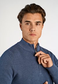 Niebieski sweter na zamek błyskawiczny z fakturowanym wzorem, wysoki kołnierz i ściągacze. Zbliżenie na dłonie trzymające zamek.