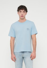 Φωτεινό μπλε βαμβακερό μπλουζάκι με κοντά μανίκια, στρογγυλή λαιμόκοψη και ένα μικρό γραφικό τύπωμα στο αριστερό στήθος. Φοριέται με φωτεινό μπλε τζιν παντελόνι.